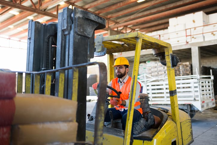 man-driving-a-forklift-wearing-a-hard-hat-and-safety-vest-moving-supplies-in-a-large-industrial-warehouse